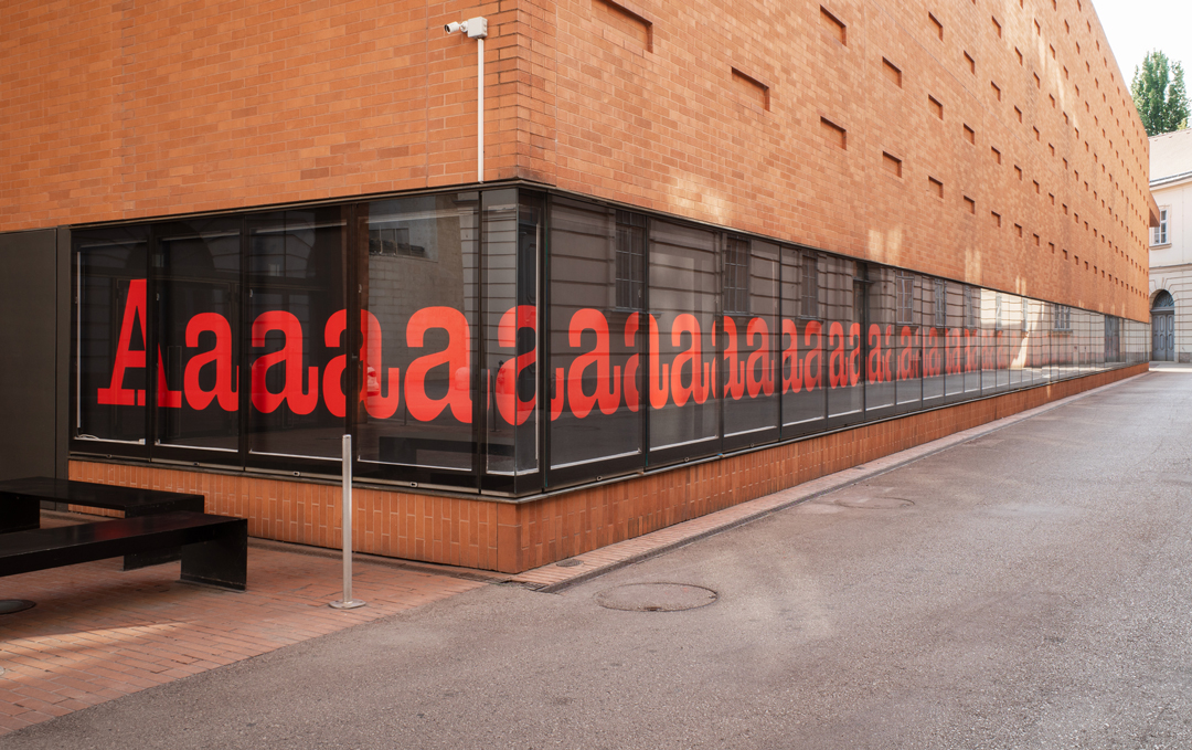 the letters 'Aaaaaaaaaaaa[…]' are painted in red on a black background. These letters are displayed inside vitrines (glass display cases) positioned on the facade of a brick building along a narrow alleyway.